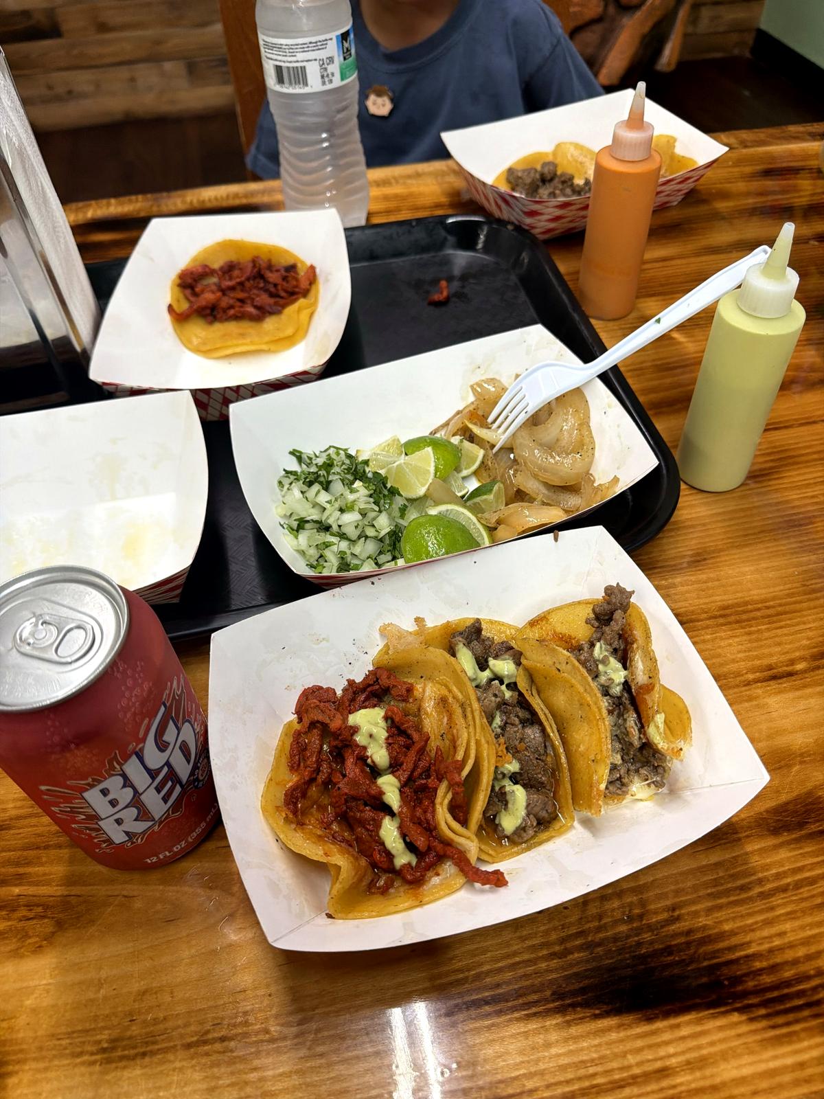 A tray of tacos in paper boats on a lacquered wood table, salsa bottles and a Big Red